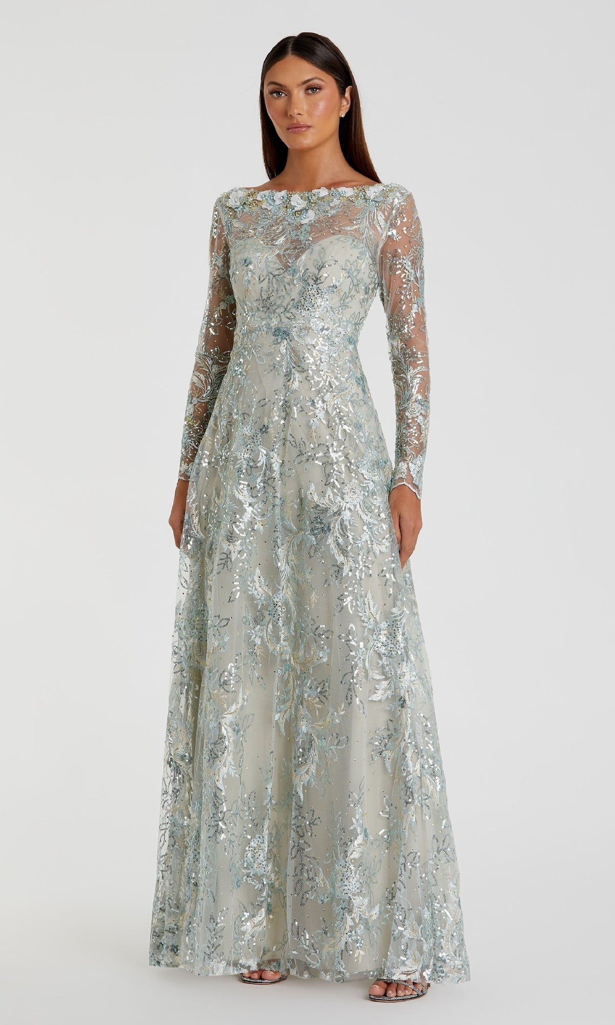 A woman stands against a plain background wearing the Mac Duggal 67498—a long formal dress with sheer long sleeves, light blue and silver floral embroidery.